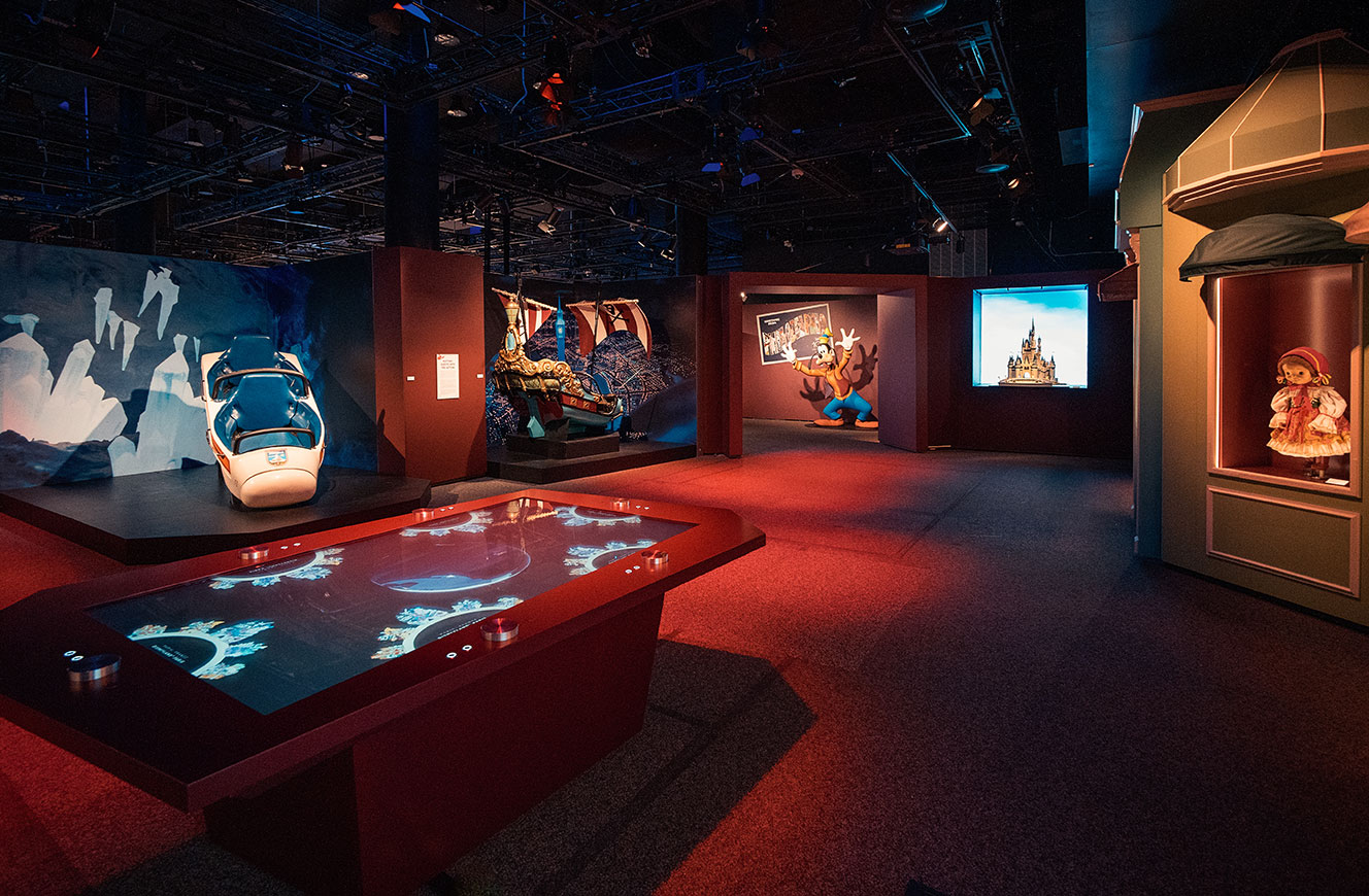 Bereich „Parks“ der Disney 100 Ausstellung mit Medientisch über internationale Disney-Themenparks, gestaltet von cocurio – ergänzt durch Karten, Modelle und Videoelemente im Hintergrund