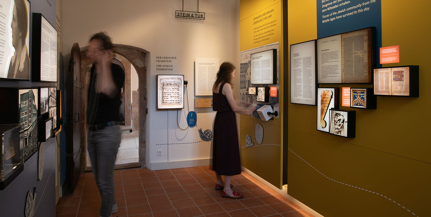 Besucher an Medienstationen im Ausstellungsraum mit gelben und grauen Wandflächen, gerahmten Exponaten und historischem Steinbogen – Stein & Tür, Gestaltung von cocurio