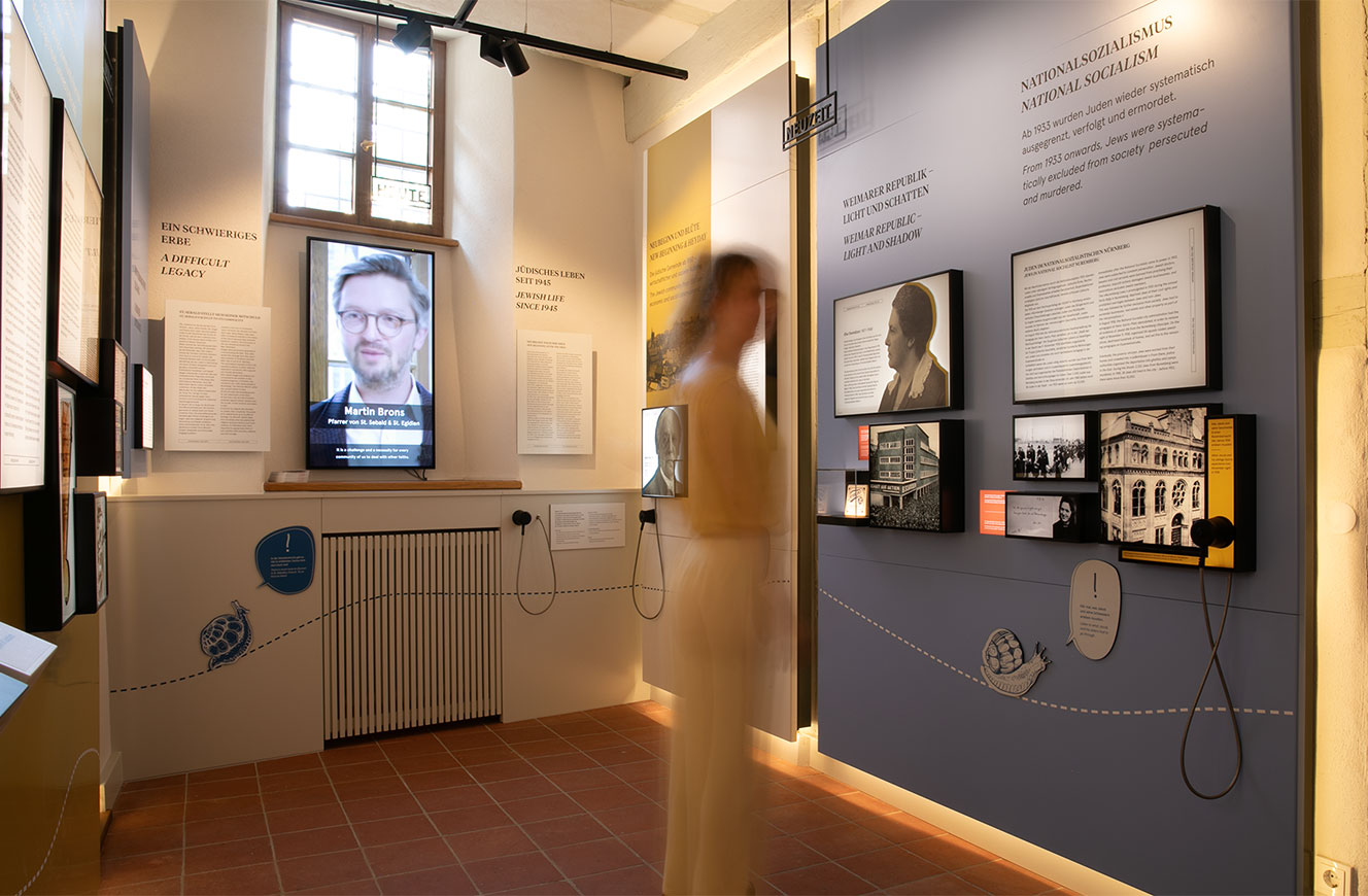 Ausstellungsraum mit Medienstation, auf dem Bildschirm ein Interview, daneben Texttafeln und historische Abbildungen – Stein & Tür