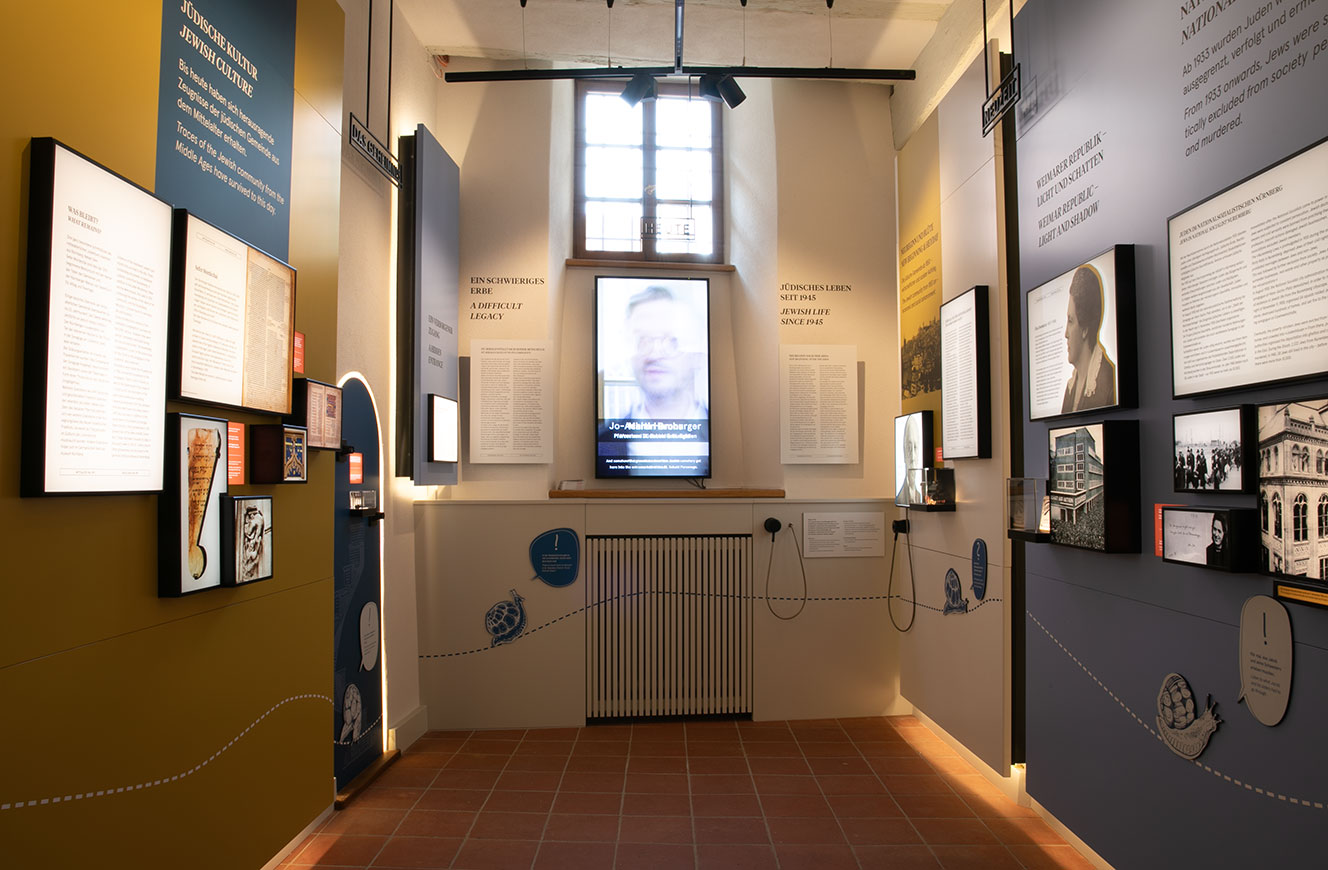 Schmaler Ausstellungsraum mit Fenster, Bildschirm und beidseitig angeordneten Text- und Bildtafeln in Blau und Gelb – Stein & Tür