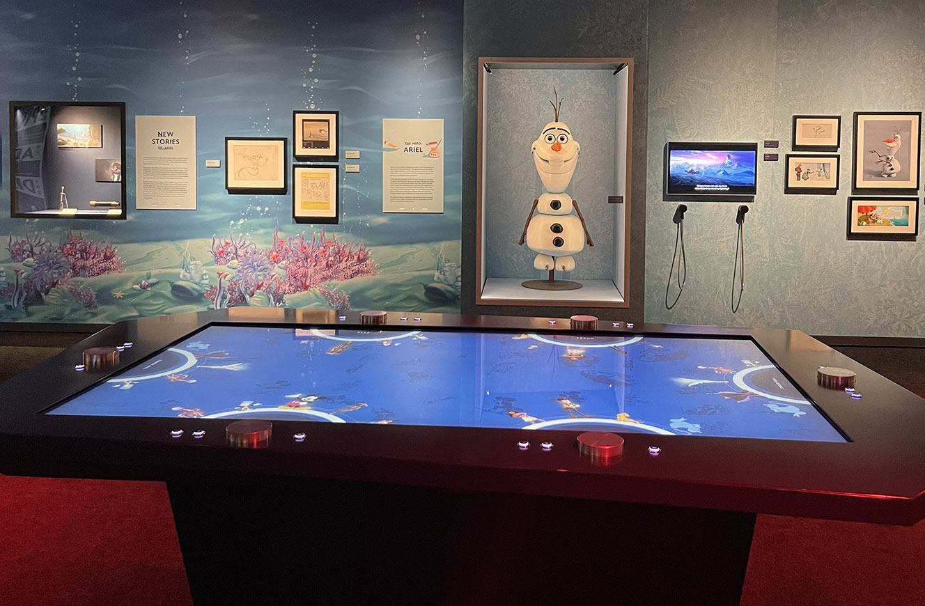 Interaktiver Medientisch mit Touch- und Drehelementen vor Original-Requisiten und Artworks zu Arielle und Olaf – Teil der Disney 100 Ausstellung, Medienkonzept und UX-Design von cocurio
