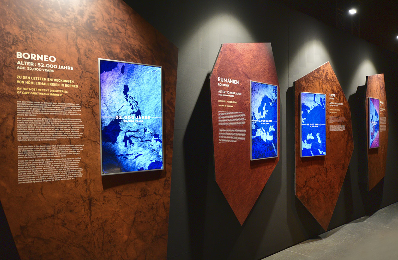 Reihe mehreckiger Infotafeln zu Höhlenfundorten in Borneo und Rumänien mit eingelassenen Bildschirmen – Lascaux