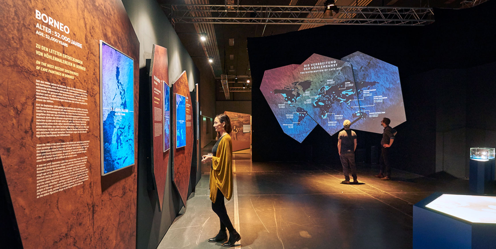 Ausstellungsraum mit großformatigen Infotafeln zu Höhlenfundorten und einer auf mehreckige Flächen projizierten Weltkarte – Lascaux