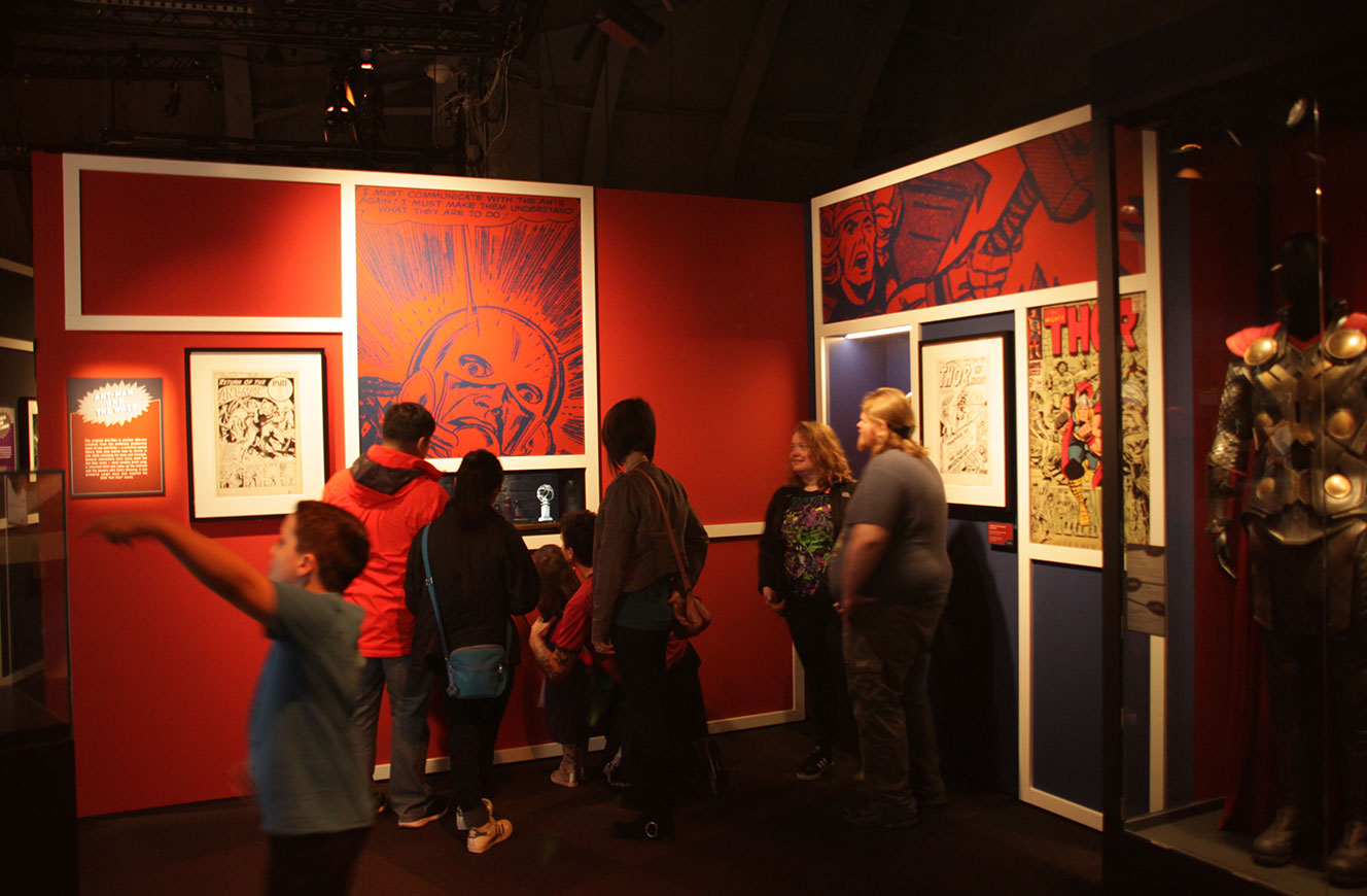 Besuchergruppe vor rot gestalteten Ausstellungswänden mit gerahmten Comicseiten und Originalzeichnungen – Marvel: Universe of Super Heroes