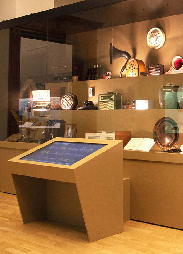 Beleuchtete Vitrine mit Radios, Uhren und Grammofontrichter, davor ein Medientisch mit Touchscreen – On Air