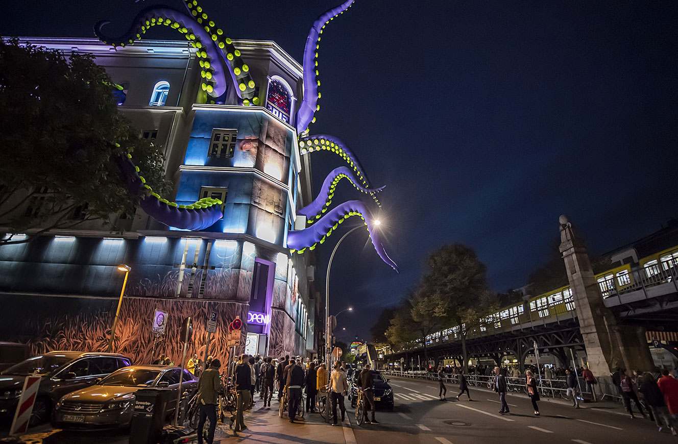 Museumsfassade bei Nacht mit riesigen violetten Tentakeln und beleuchteter Wandmalerei, davor Besucher auf der Straße – Urban Nation Biennale