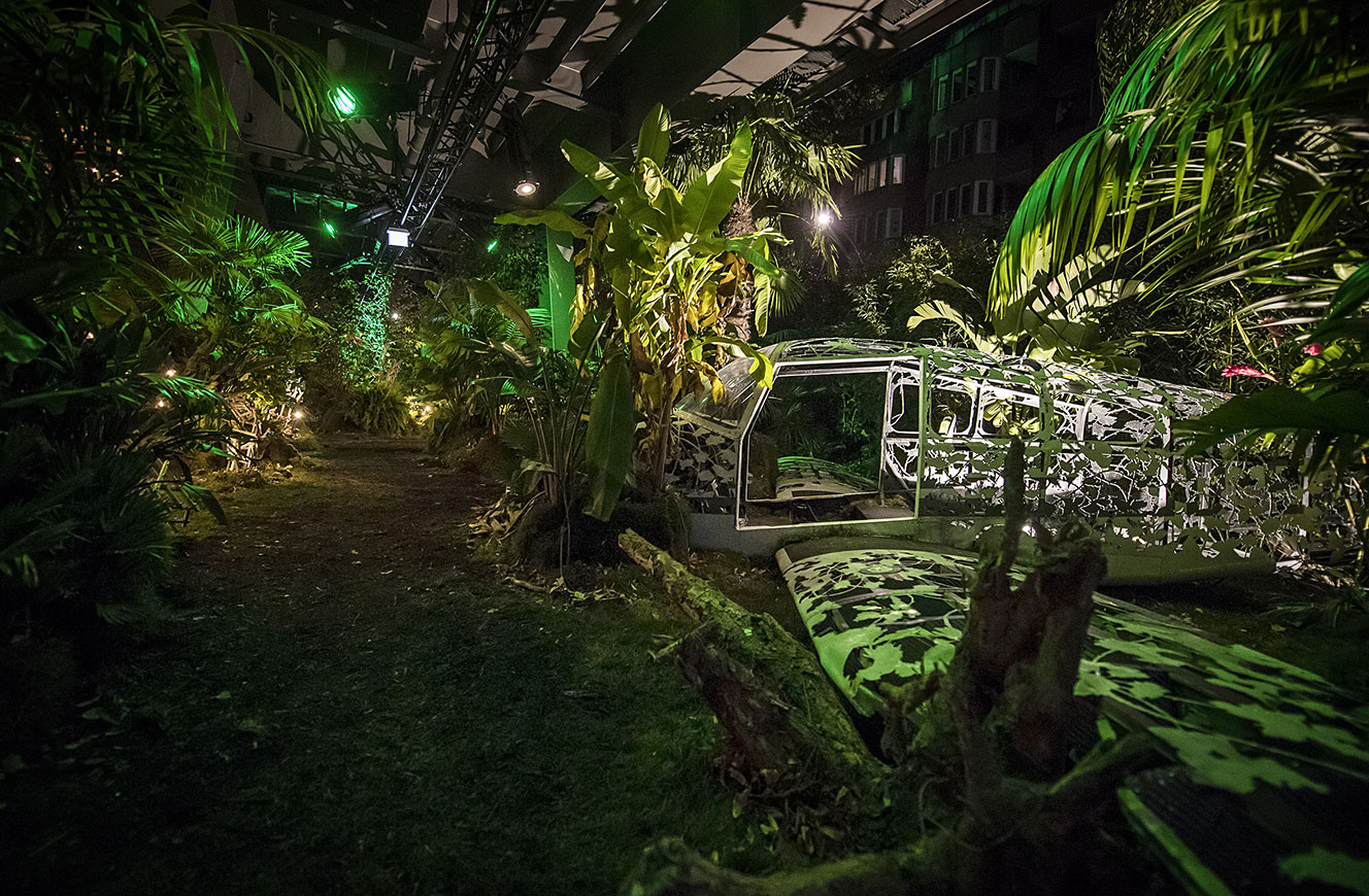 Nächtliche Dschungel-Installation mit dichter Bepflanzung, farbigem Licht und einem überwucherten Fahrzeug – Urban Nation Biennale