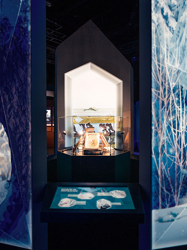 Beleuchtete Vitrine mit historischen Büchern und Zeichnungen, davor die Medienstation „Sources of Inspiration“ – Disney 100