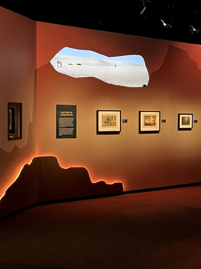 Ausstellungswand in Rot mit projizierter Wüstenlandschaft, gerahmten Zeichnungen und der Texttafel „Nature in Animation“ – Disney 100